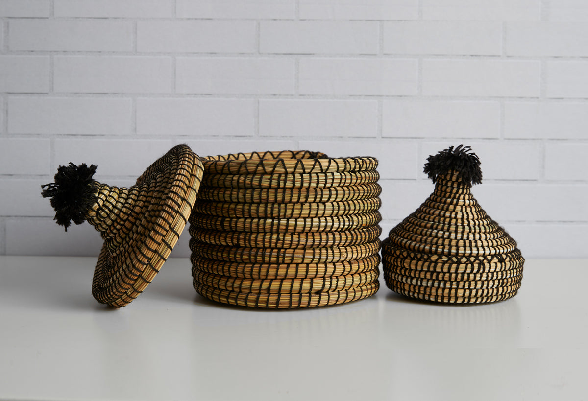 Two woven straw baskets with black tassels, one cylindrical and one round with pointed lid.
