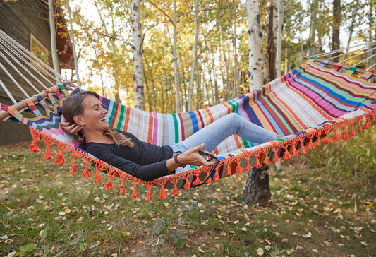 Colorful striped loomed hammock with orange fringe hung between trees, person reclining inside.