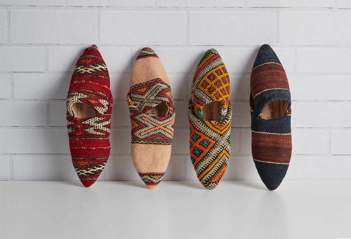 Four kilim slippers with geometric patterns in red, beige, gold, and navy, displayed from above against white background.