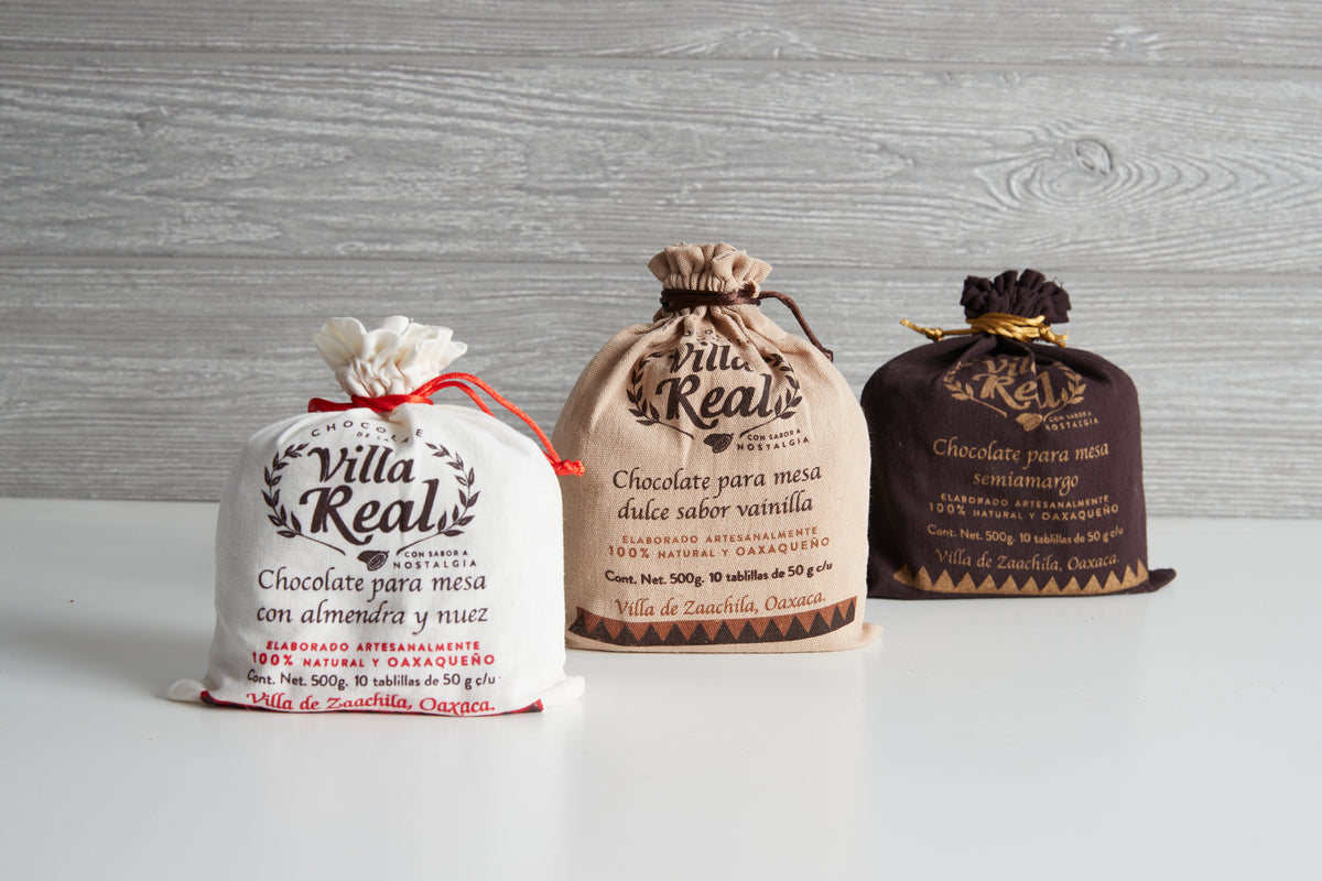Three Villa Real Mexican hot chocolate packages with drawstring closures in cream, tan, and dark brown colors on gray wood ba