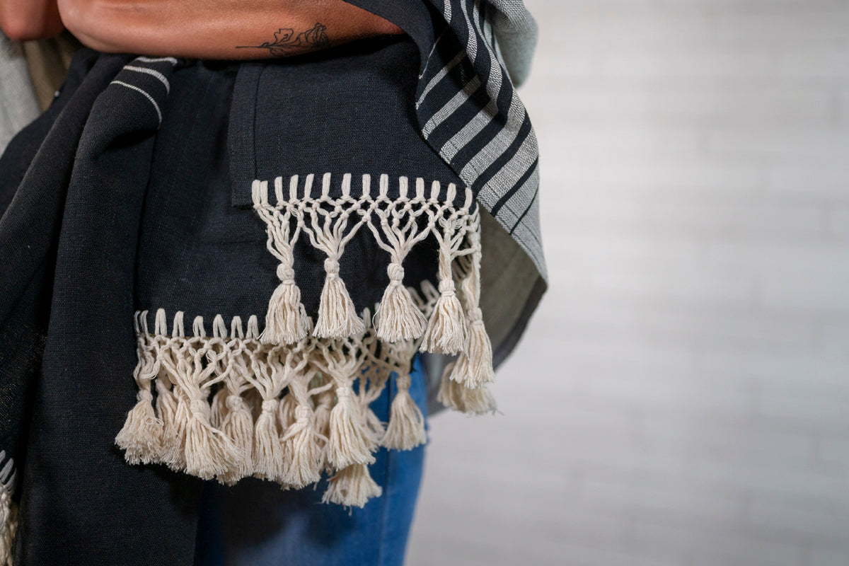 Black woven blanket with cream knotted fringe trim, draped over person's shoulder.