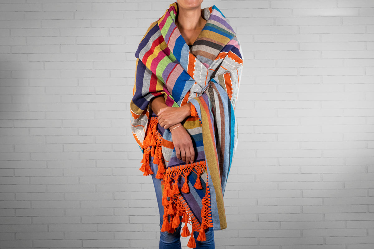 Colorful striped blanket with orange tassels worn draped over a person's shoulders against a white brick wall.