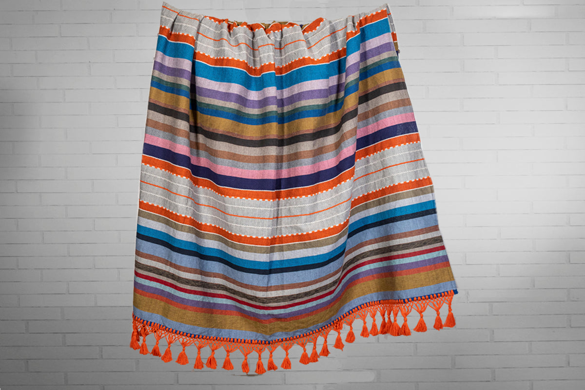 Woven blanket with horizontal stripes in orange, blue, purple, gold, and cream, trimmed with orange tassel fringe.