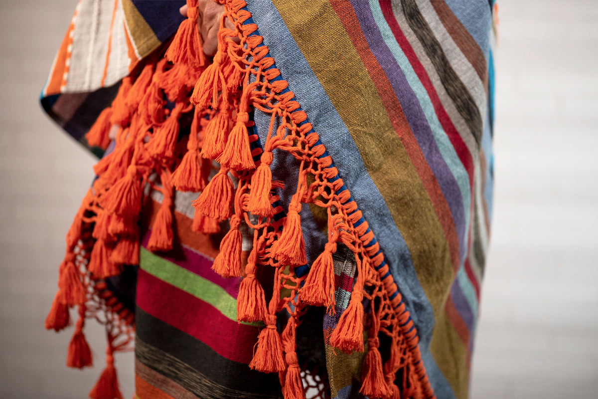 Woven blanket with multicolored horizontal stripes and orange tassel fringe trim along the edge.