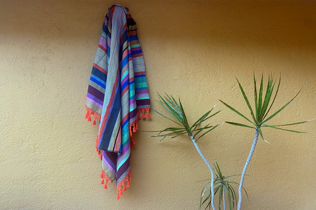 Striped textile blanket with multicolor horizontal bands and coral fringe trim draped against a yellow wall.