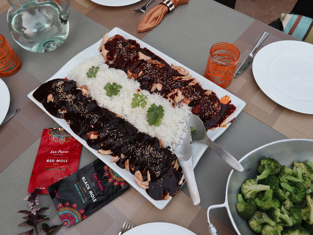 Enchiladas covered in dark mole sauce, white rice, and cilantro on white plate with Los Pacos mole packets nearby.