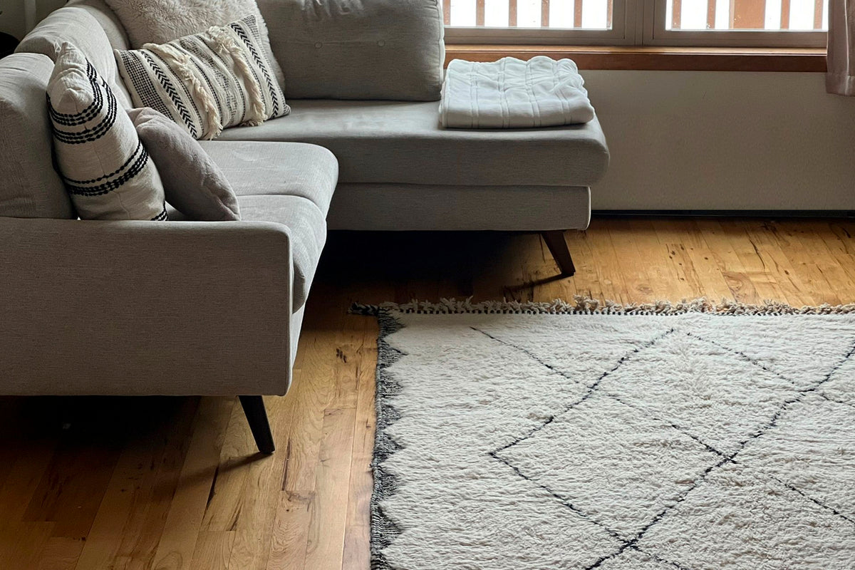 Cream wool rug with dark diamond pattern displayed on wooden floor in front of gray sectional sofa.