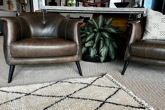 Cream wool rug with black geometric diamond pattern placed in living room with leather chairs and potted plant.