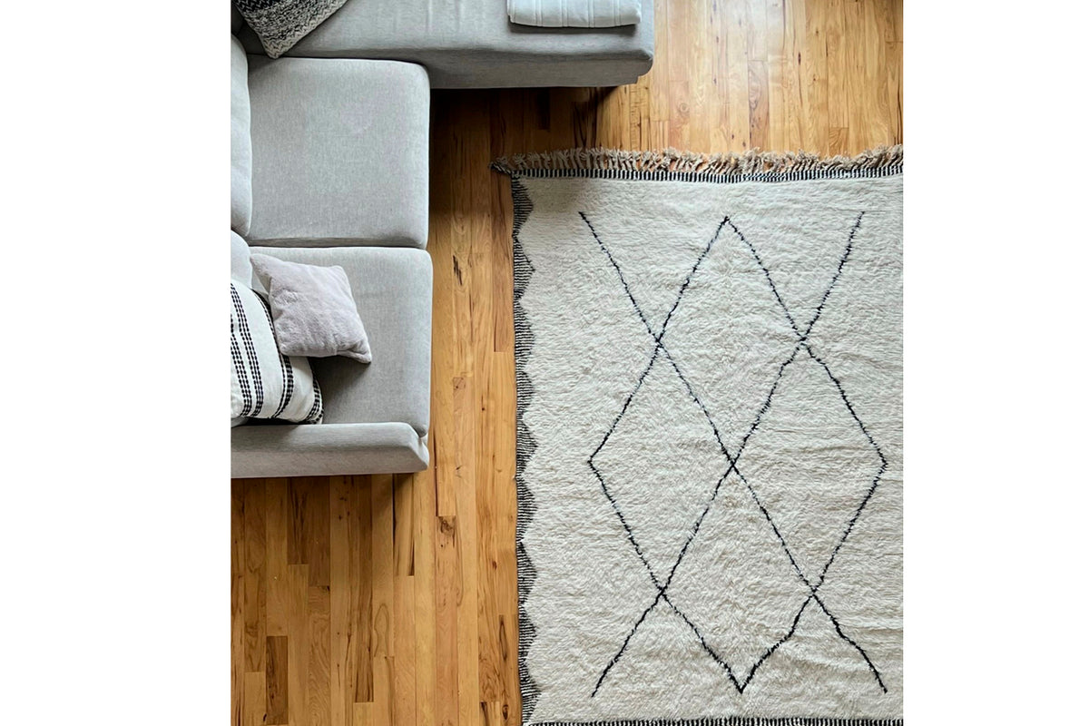 Cream wool rug with black diamond pattern on wooden floor next to gray sectional sofa.