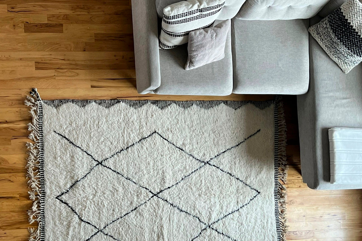 Cream-colored wool rug with black diamond pattern and fringe edges, displayed on hardwood floor next to gray furniture.