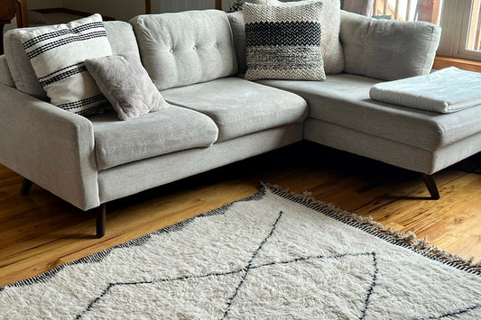 Cream-colored wool rug with black diamond pattern and fringe edges displayed on wooden floor in front of light gray sectional