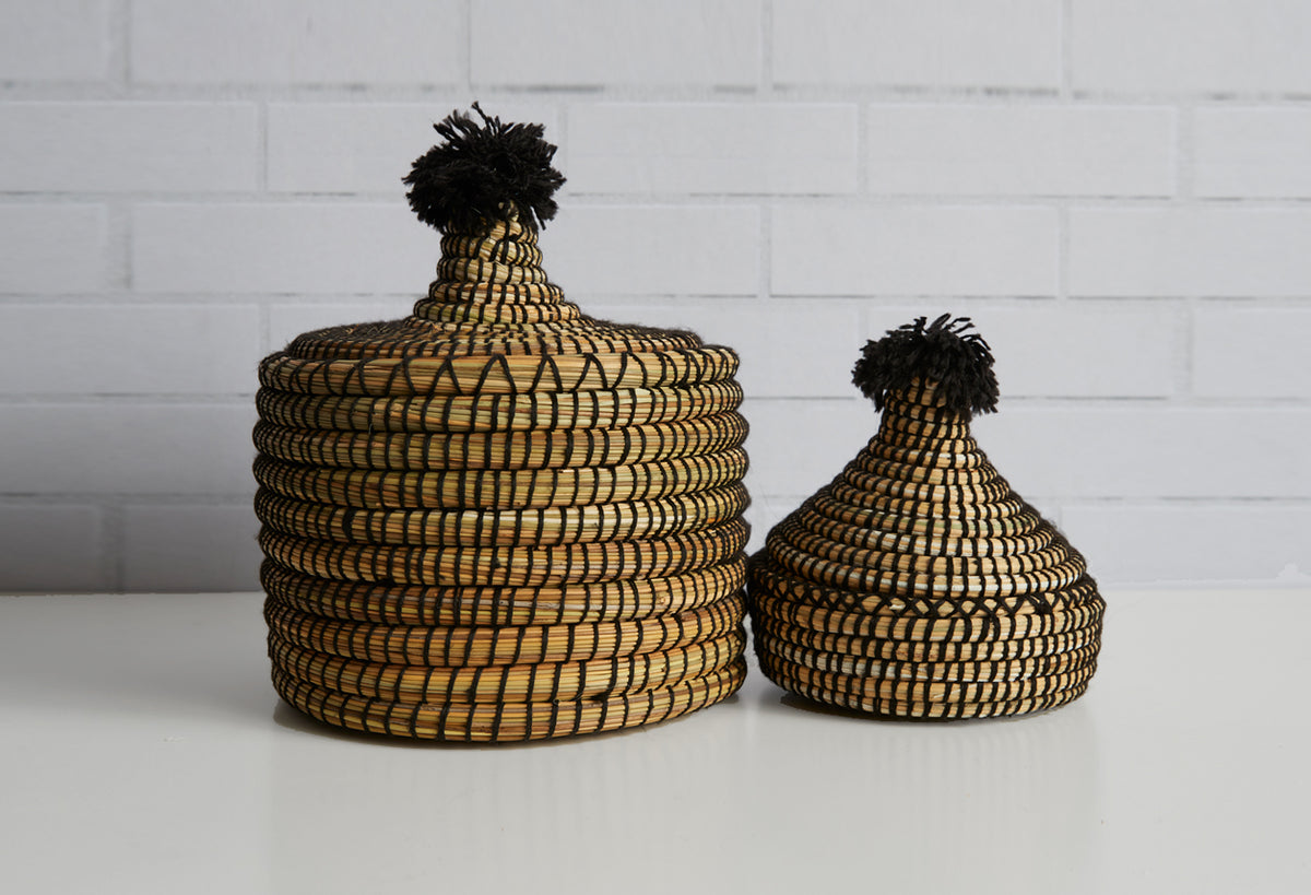 Two woven baskets with black pom-poms on lids, one larger and one smaller, in tan and black pattern.
