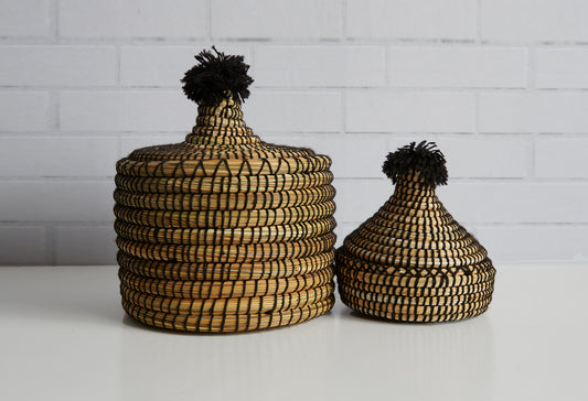 Two woven baskets with black pom-poms on lids, one larger and one smaller, in tan and black pattern.