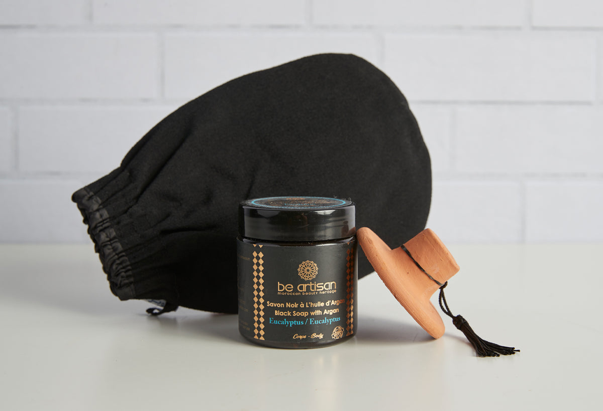 Black exfoliating glove, wooden massage brush, and black soap jar with gold labeling displayed together.