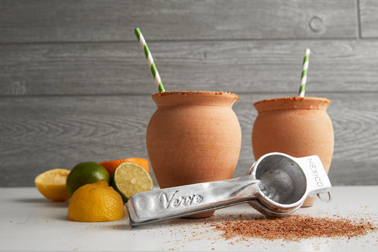 Two terracotta cups with green straws, metal measuring spoons, lemons, lime, and cinnamon powder on white surface.