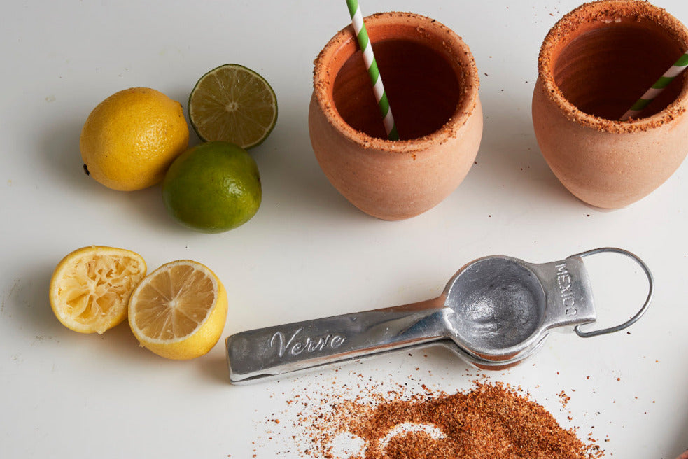 Two clay cantaritos with striped straws, fresh lemons and limes, metal measuring spoon, and chili powder seasoning.
