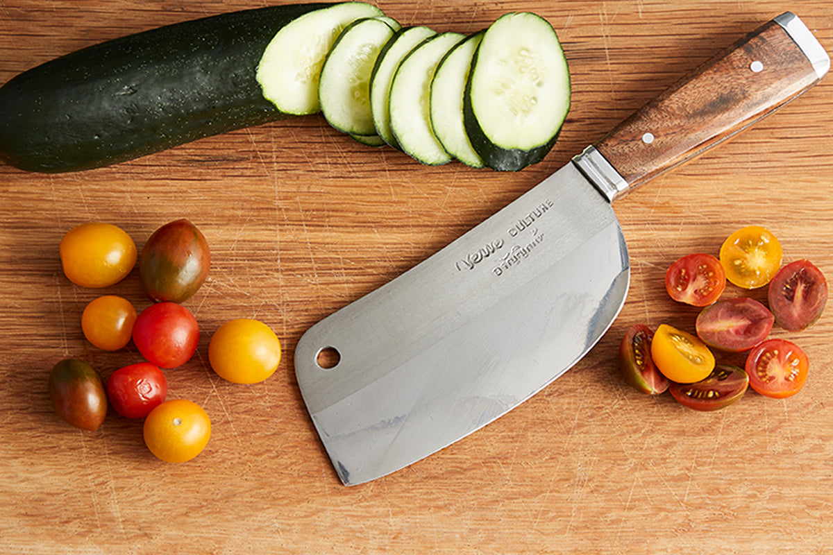 Thai chef's knife with wooden handle displayed on cutting board surrounded by sliced cucumber and tomatoes.