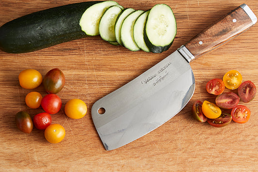 Thai chef's knife with wooden handle displayed on cutting board surrounded by sliced cucumber and tomatoes.