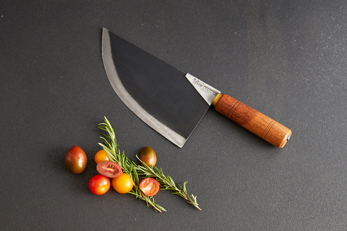 Curved chef's knife with wooden handle displayed above fresh tomatoes and rosemary sprigs on gray surface.