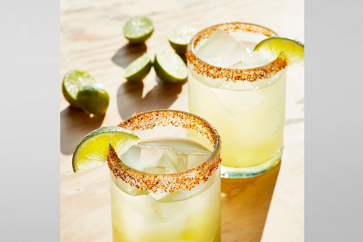 Two clear handblown glasses filled with yellow-green beverage, lime slices, and ice with spiced rims.