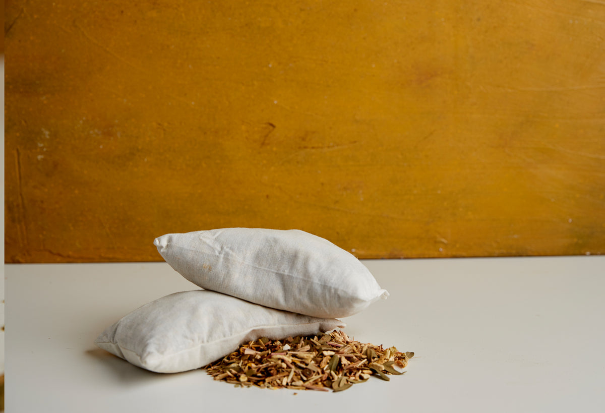 Two white cloth pouches and pile of dried herbs on white surface against mustard-yellow background.