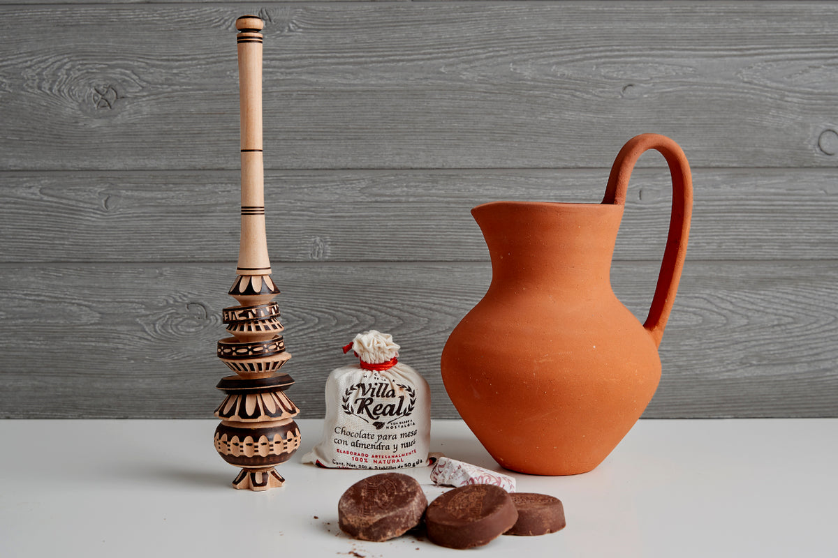 Wooden molinillo whisk, terracotta pitcher, packaged hot chocolate mix, and chocolate tablets arranged on white surface again