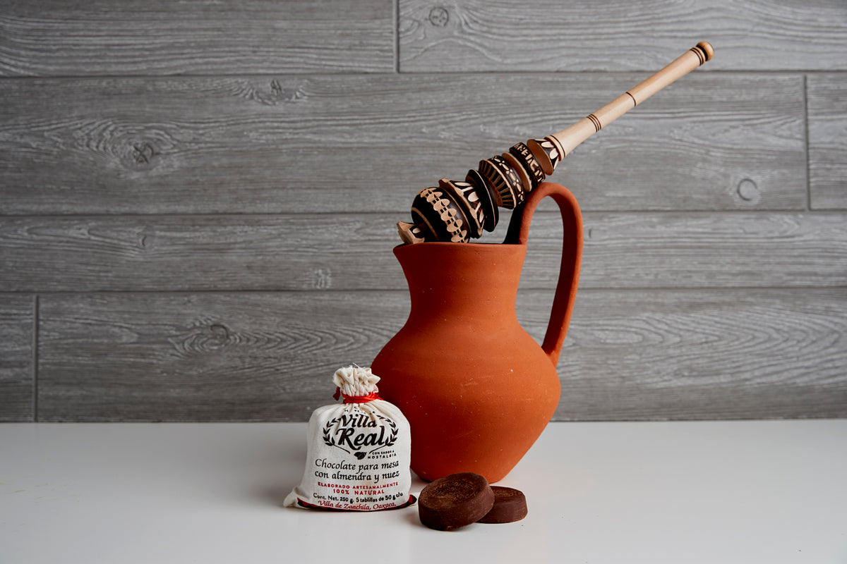 Terracotta pitcher with wooden molinillo, packaged chocolate tablet, and chocolate discs on gray wood background.