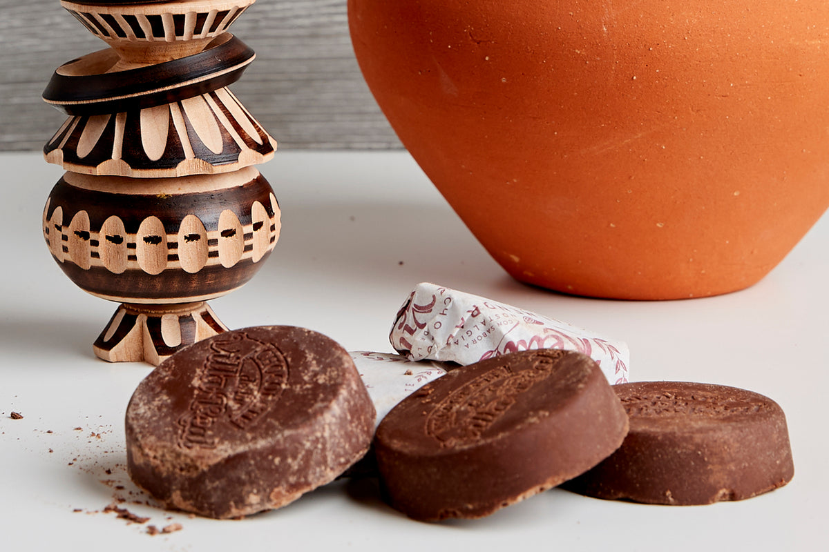 Wooden whisk with decorative carved patterns, orange ceramic bowl, and three chocolate discs with textured surfaces.