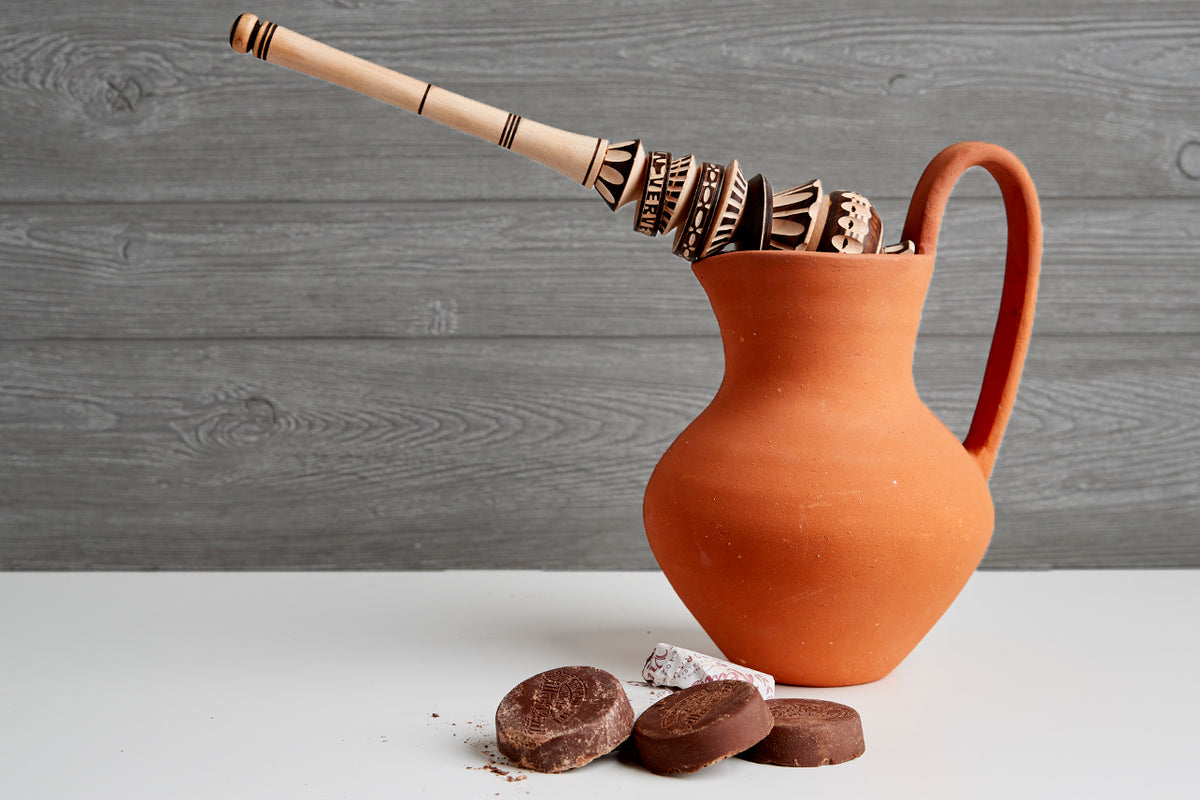 Terracotta jug with red handle, wooden stirrer with decorative bands, and chocolate truffles displayed in front.