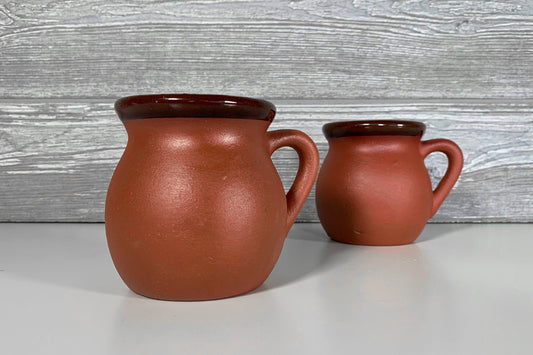 Two terracotta mugs with handles and dark brown rims in different sizes.