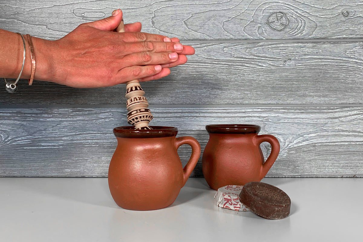 Two terracotta mugs with handles and wooden lids, displayed with chocolate and a decorative spoon.