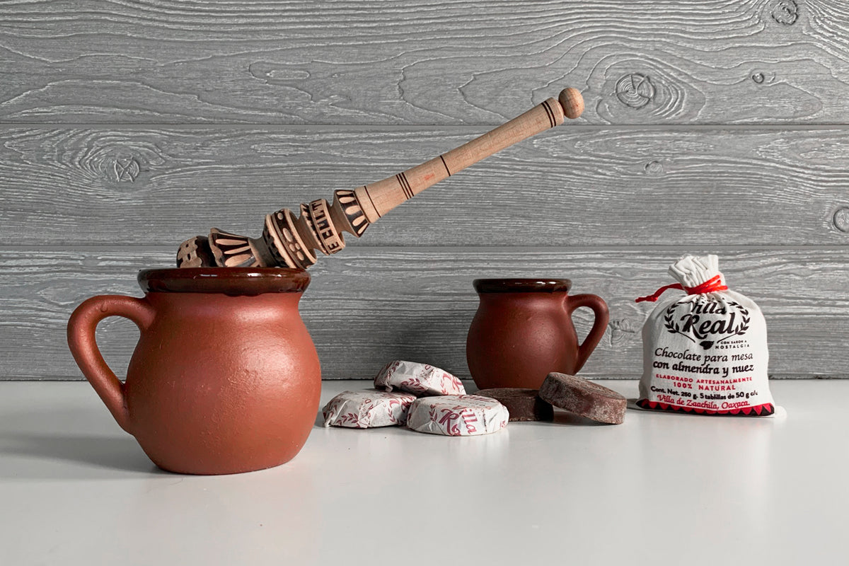 Terracotta pot with wooden whisk, smaller ceramic pitcher, chocolate tablets, and packaged mix on gray wooden surface.
