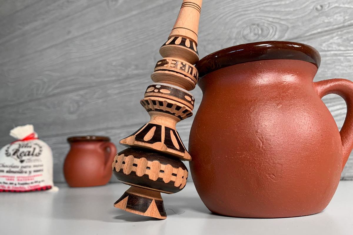 Terracotta pitcher with wooden molinillo whisk and small fabric pouch containing hot chocolate mix.