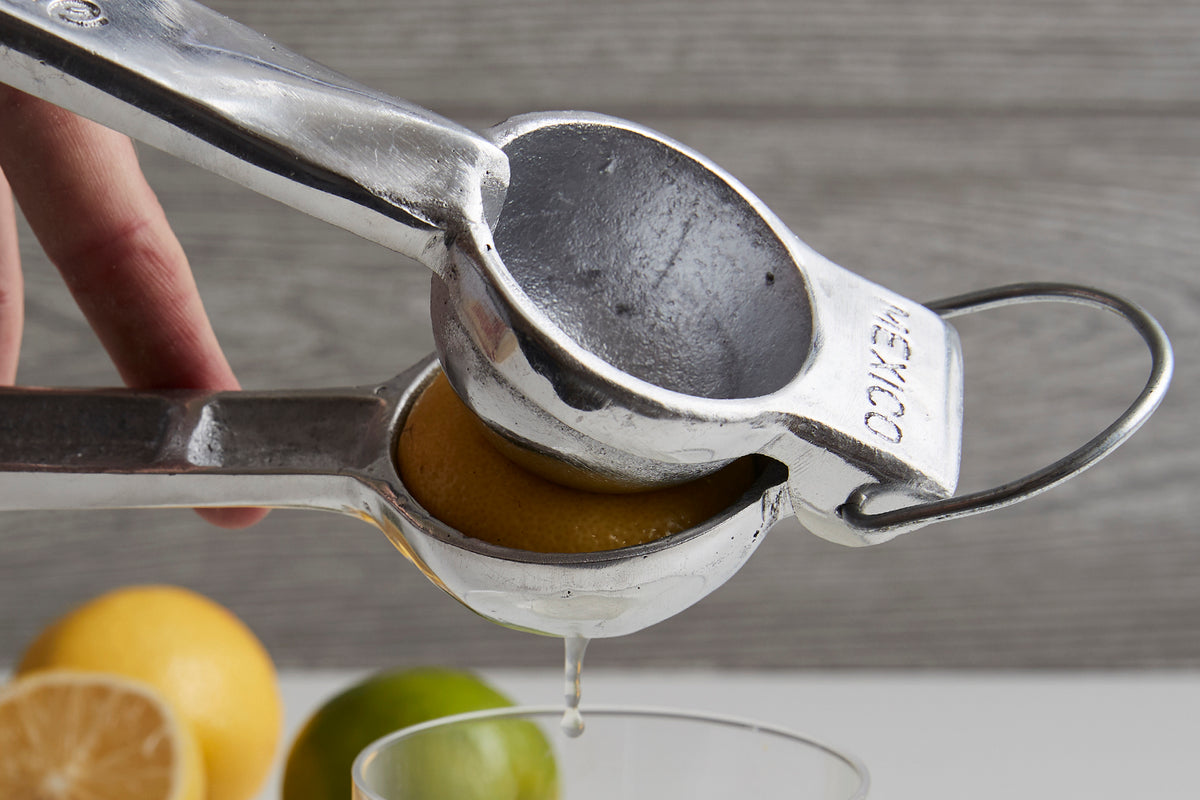 Aluminum hand juicer with two handles squeezing juice from a lemon half into a bowl, with fresh citrus fruit visible.