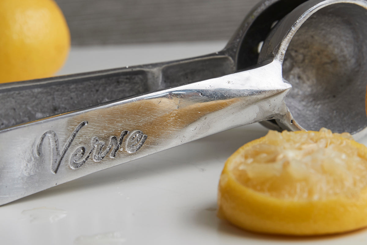 Metal hand juicer with two circular cups and handle stamped with "Vomilo," displayed with lemons and juice.
