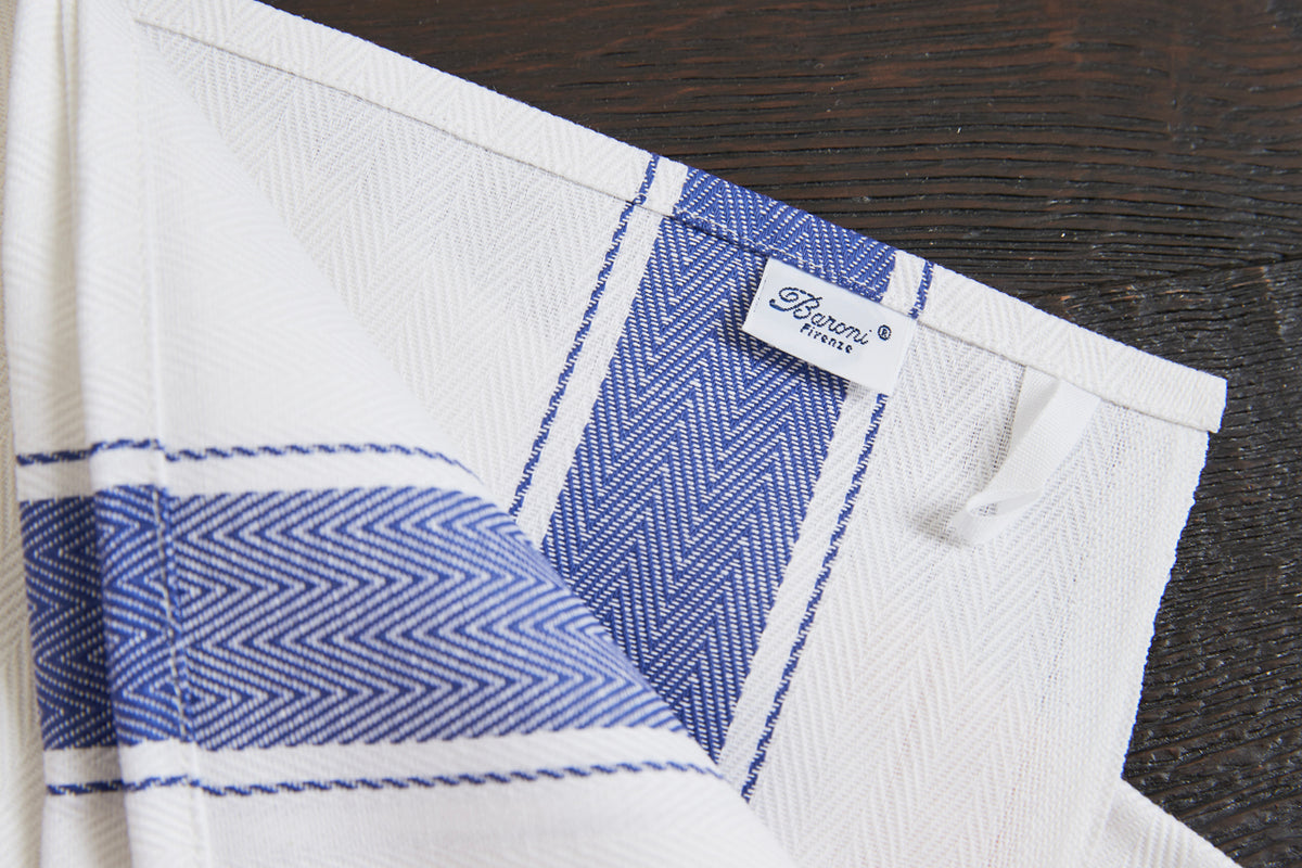 White kitchen towel with blue herringbone pattern stripe and brand label, laid on dark wood surface.