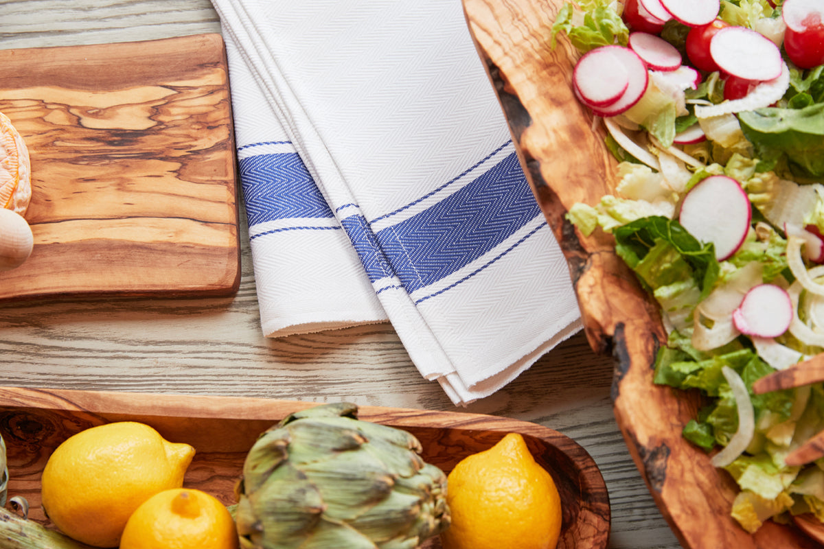 White kitchen towel with blue herringbone stripe and border, displayed on wooden surface with fresh vegetables.