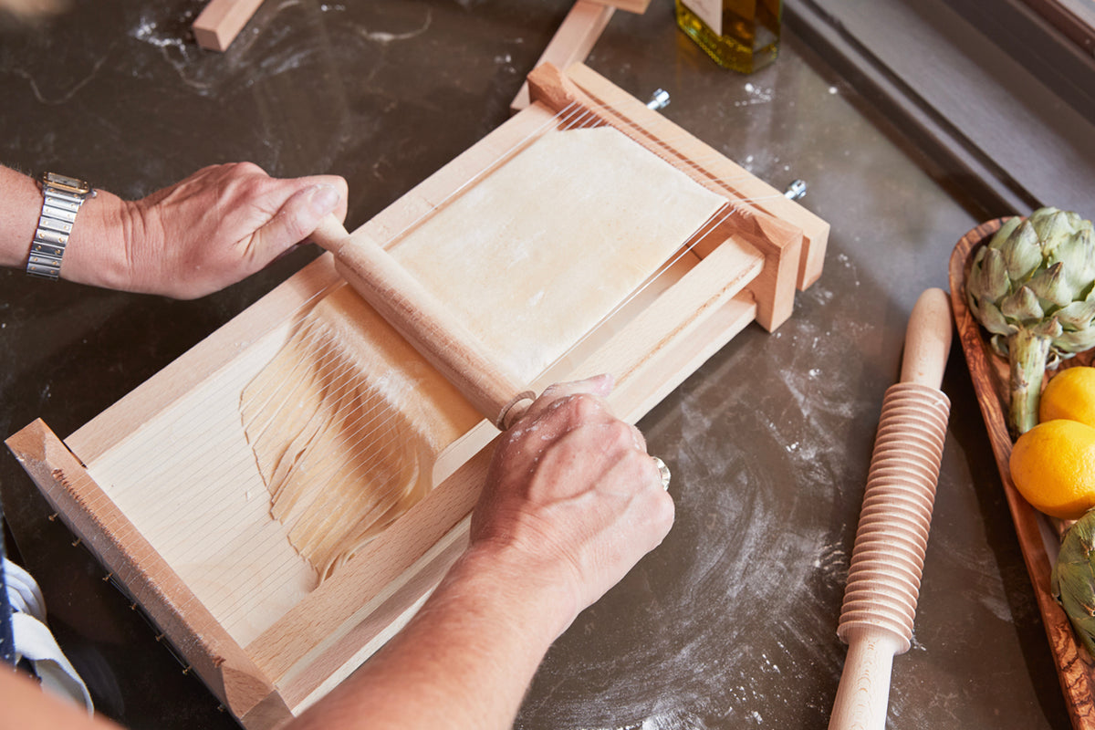 Wooden pasta maker with parallel strings and matching rolling pin on flour-dusted work surface.