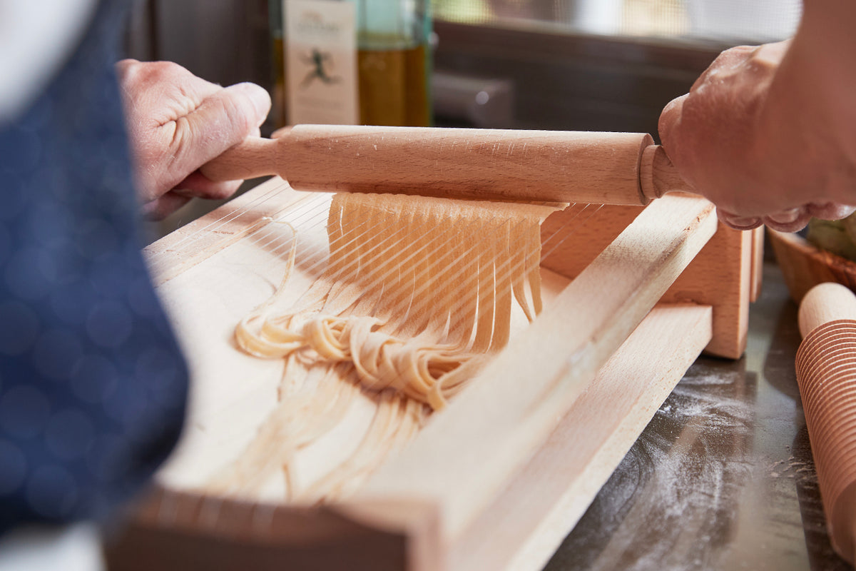 Hands using wooden rolling pin on pasta chitarra with freshly cut strands of pasta.