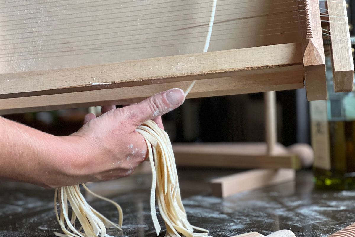 Wooden pasta maker with parallel strings producing fresh pasta strands held in hand.