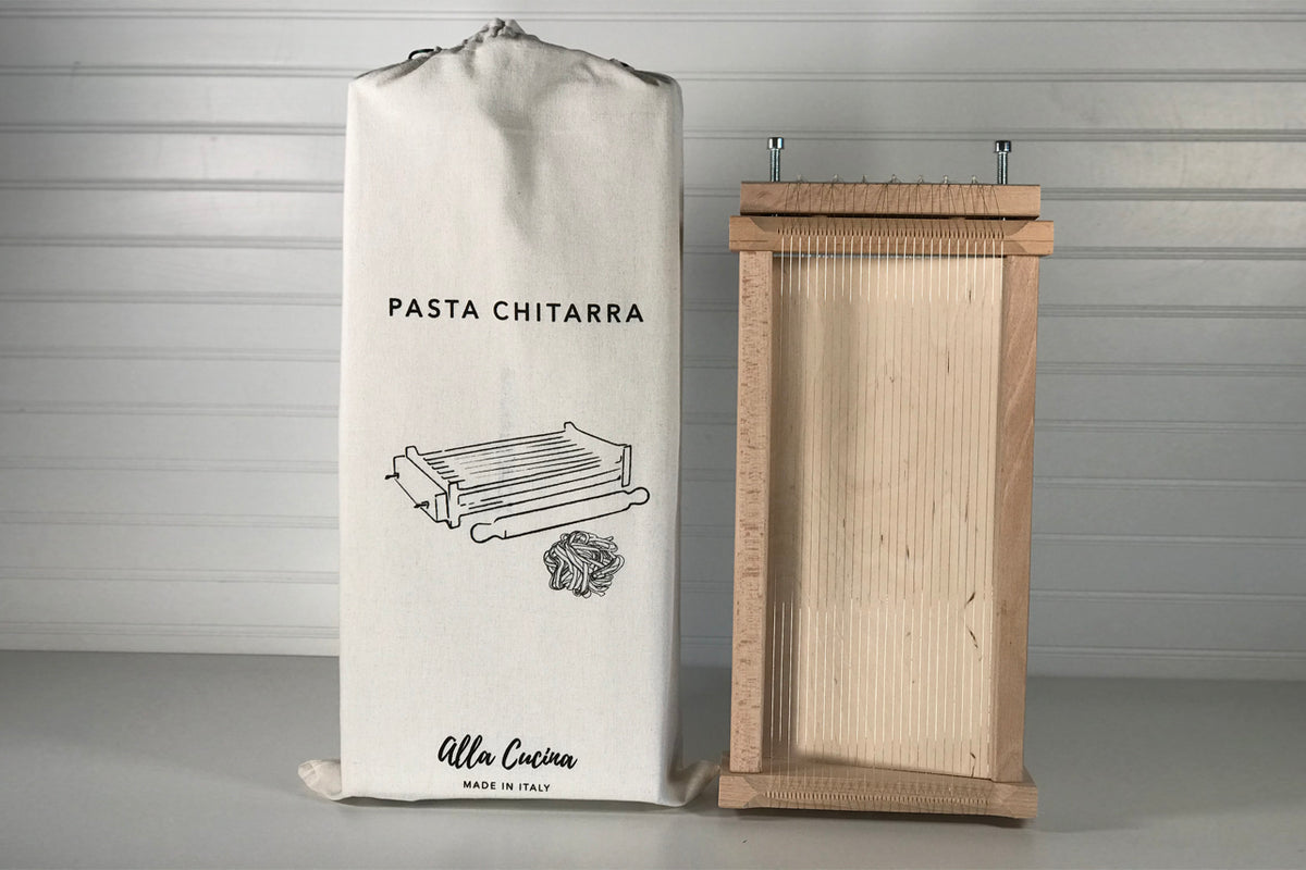Wooden pasta chitarra with parallel strings and wooden rolling pin, displayed with cream canvas storage bag.