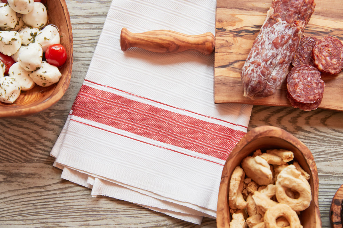 White kitchen towel with red herringbone stripe pattern and red trim lines, displayed on wooden surface with food items.