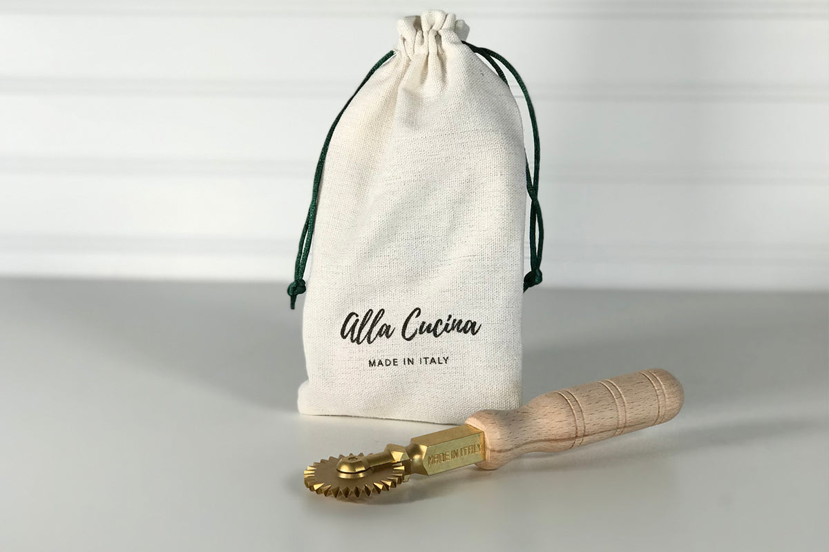 Brass fluted wheel tool with wooden handle next to cream drawstring storage bag labeled "alla Cucina MADE IN ITALY."
