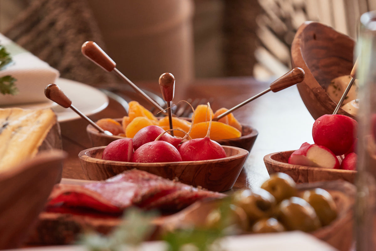 Four wooden bowls with olive wood grain holding red apples, orange citrus slices, and green olives with metal pick utensils.