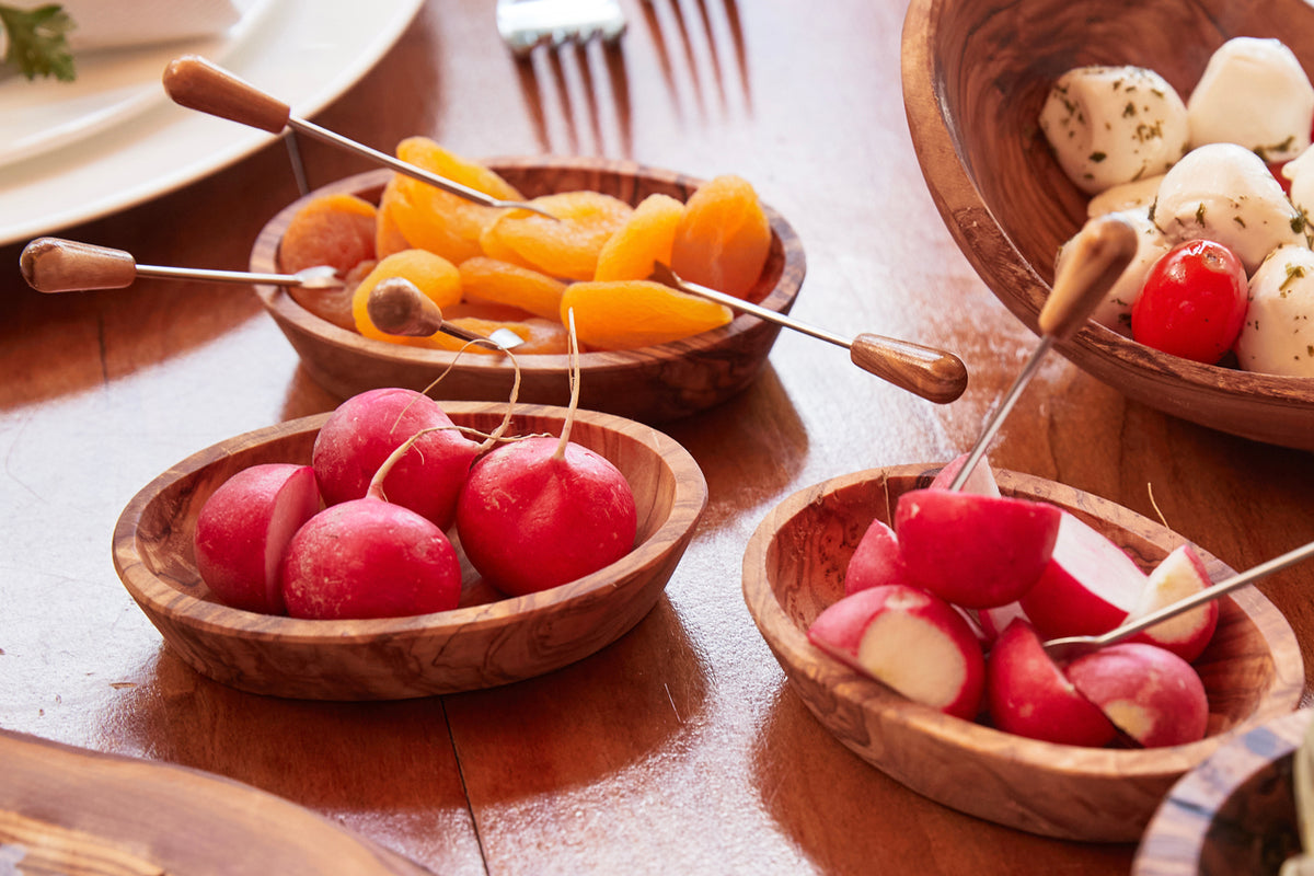 Six small forks with wooden handles displayed in olive wood bowls containing fresh vegetables and fruits.