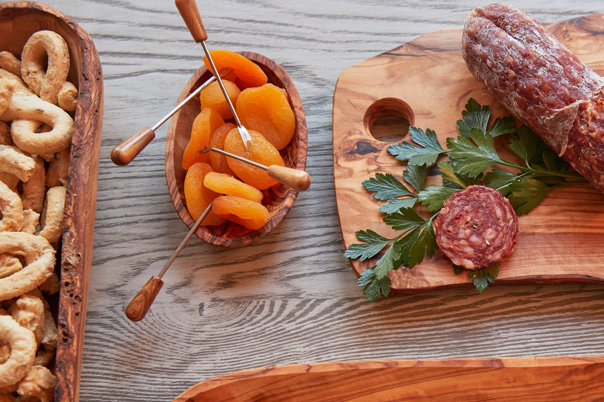 Six small forks with wooden handles displayed with appetizers including dried apricots, crackers, and cured meats on wooden s