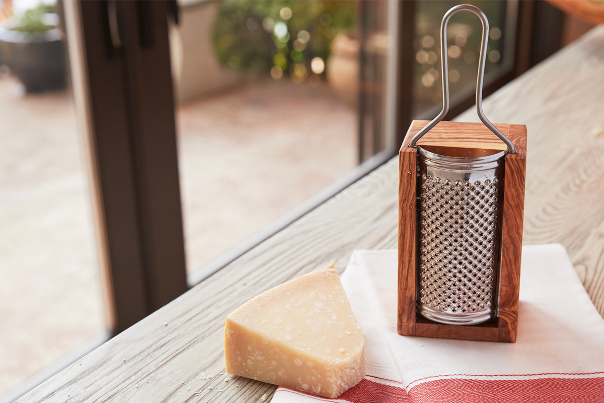 Box grater with olivewood frame, metal handle, and cylindrical grating drum next to wedge of Parmesan cheese.