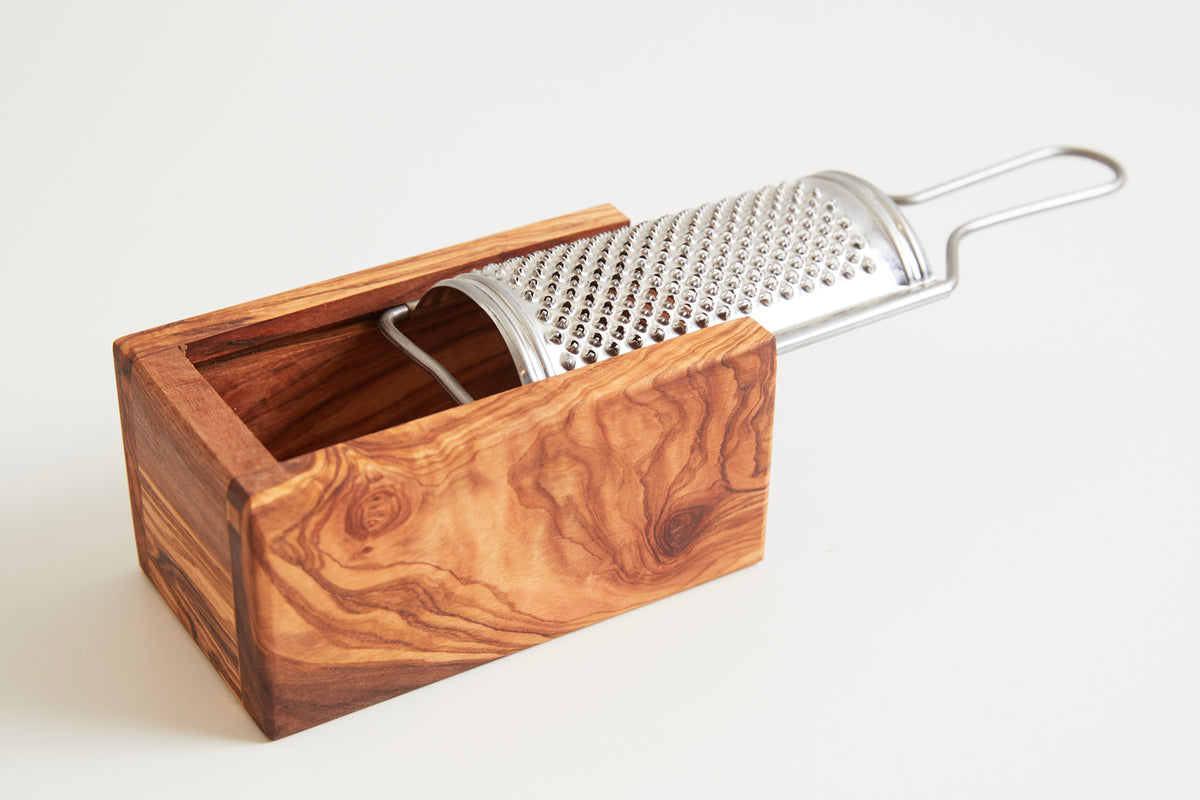 Cylindrical metal grater with stainless steel handle resting in olive wood box with open top.