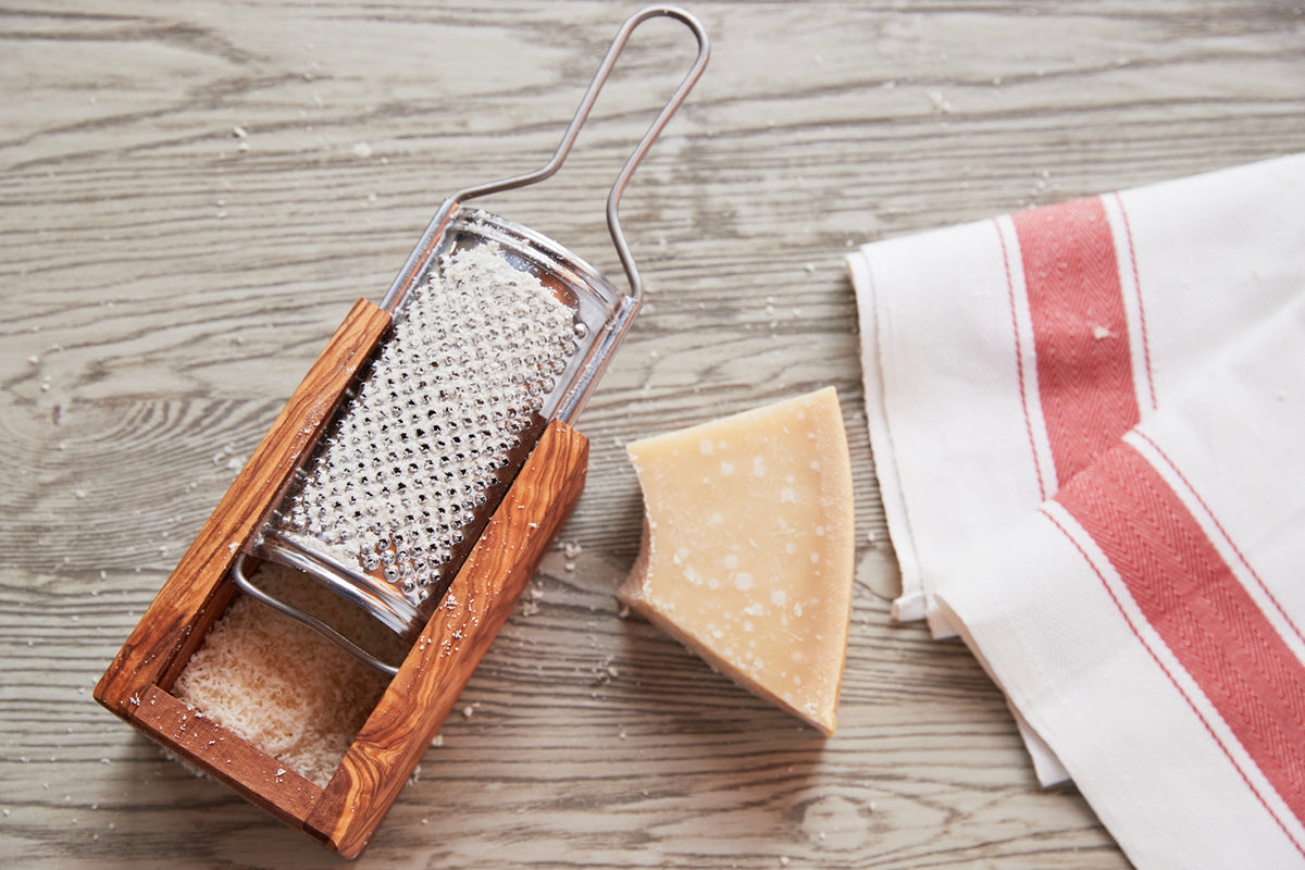 Box grater with olivewood base and metal grating surface, displayed with Parmesan cheese wedge and striped kitchen towel on w