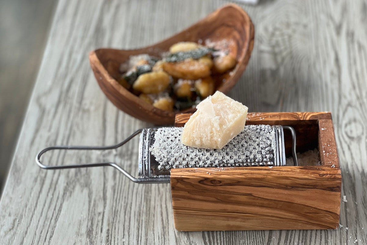 Wooden box grater with metal grating surface and handle, holding grated cheese, with olives and bread in background.
